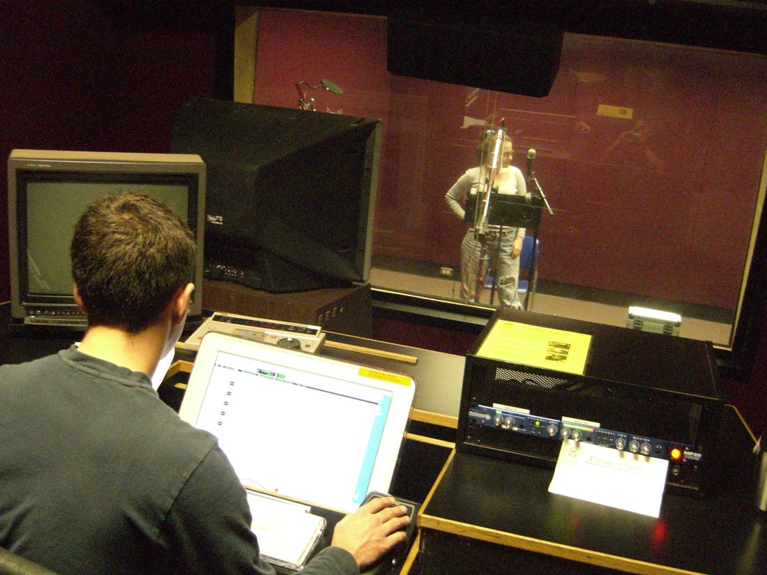 An engineer at a mixing desk records a performer in a sound booth